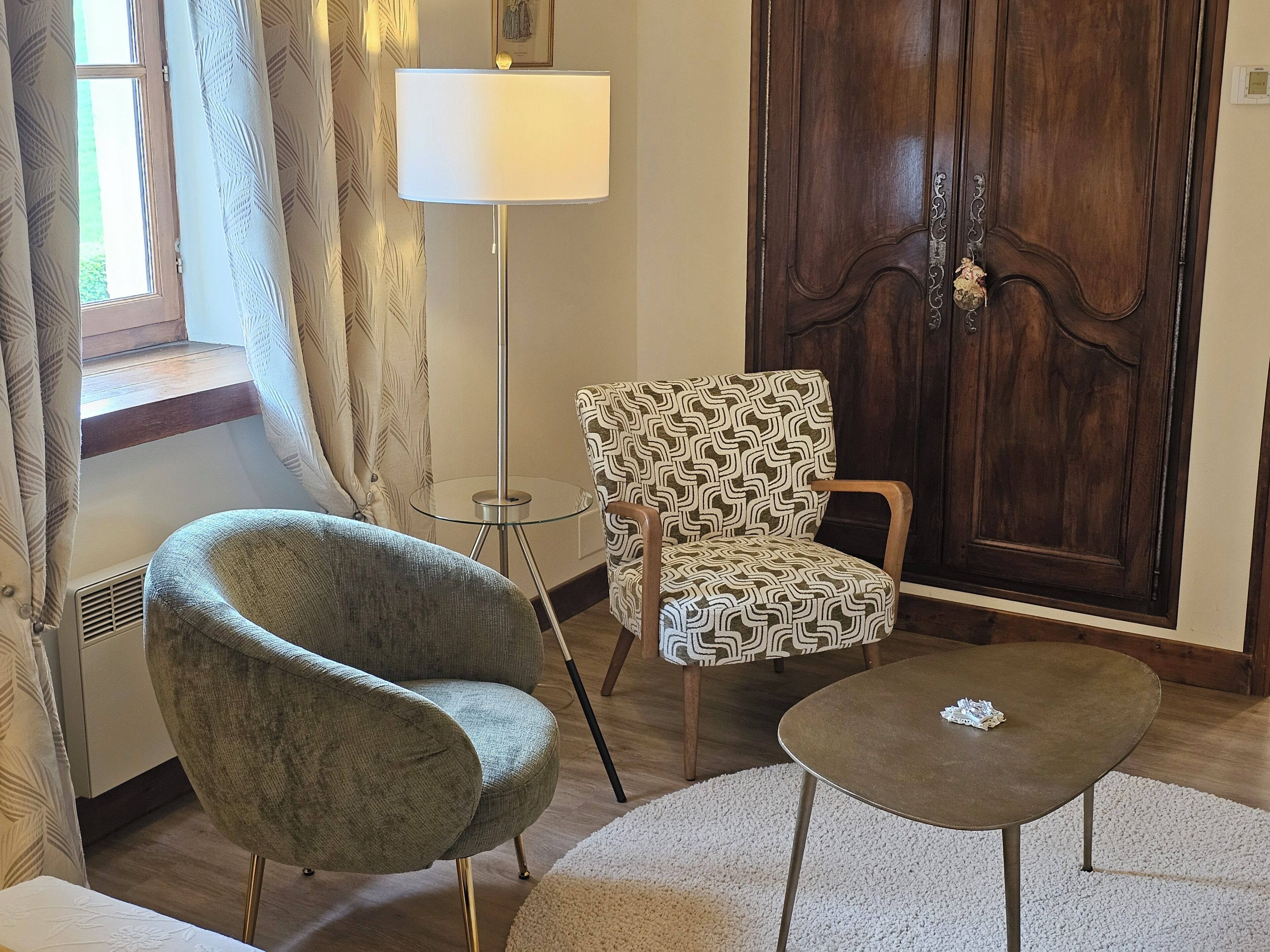 Una habitación con una puerta de madera, un sillón estampado, un sillón gris, una mesita redonda pequeña y una lámpara de pie.