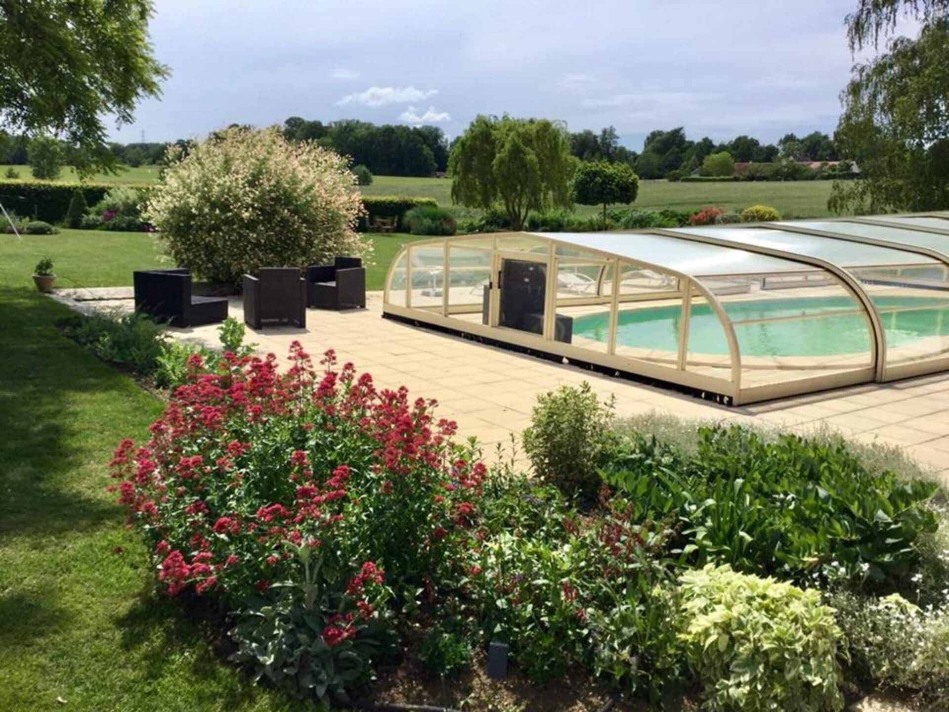 Ein überdachter Pool mit Glasveranda, umgeben von einem Garten mit verschiedenen Pflanzen und Blumen.