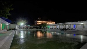 Vista nocturna de un patio de hotel con piscina, un edificio grande con iluminación cálida y una valla blanca que rodea el área.