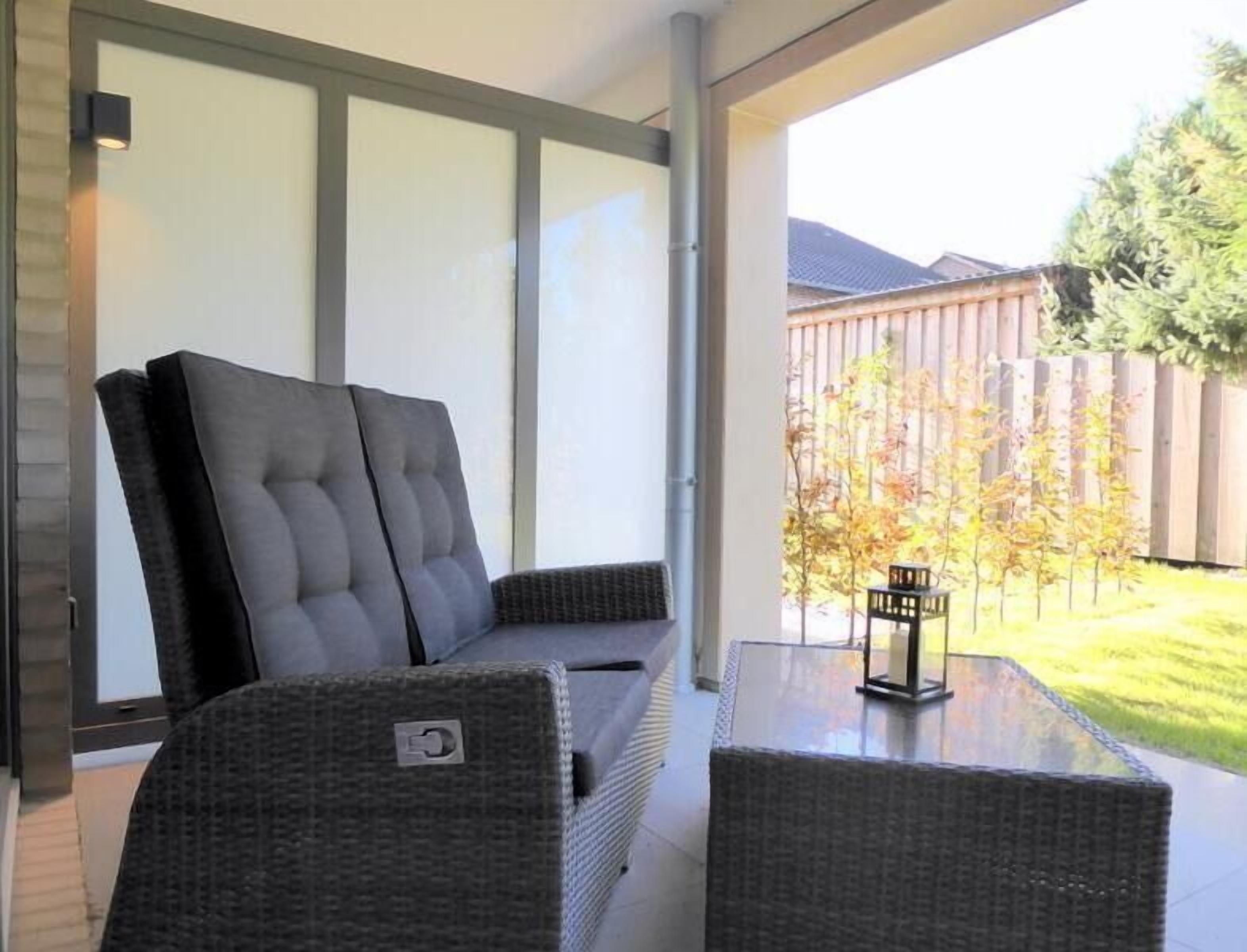 Un espace patio aménagé avec un canapé et deux fauteuils, une table en verre, et une vue sur un jardin avec des arbres et une clôture en bois.