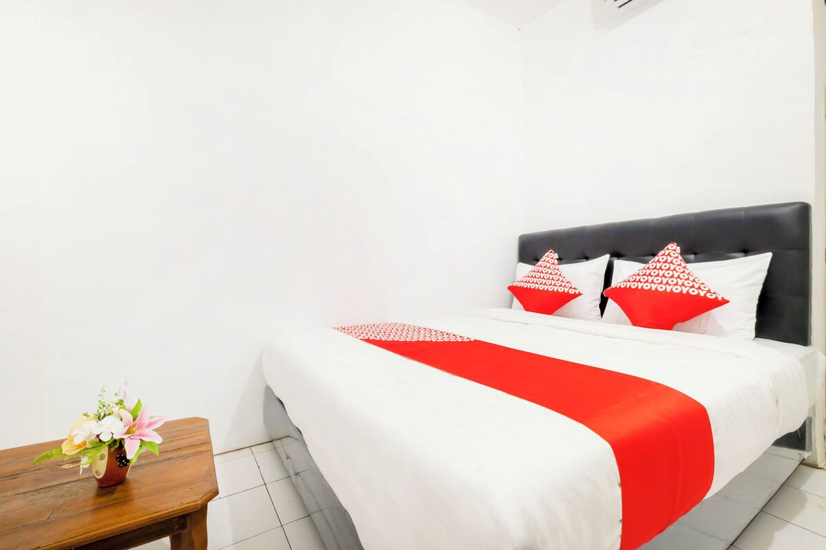 A hotel room with a bed, a nightstand with a vase, and a white wall.