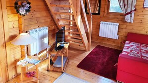 A wooden cabin with a wooden staircase, a red sofa, a small table with a lamp, and a window with curtains.