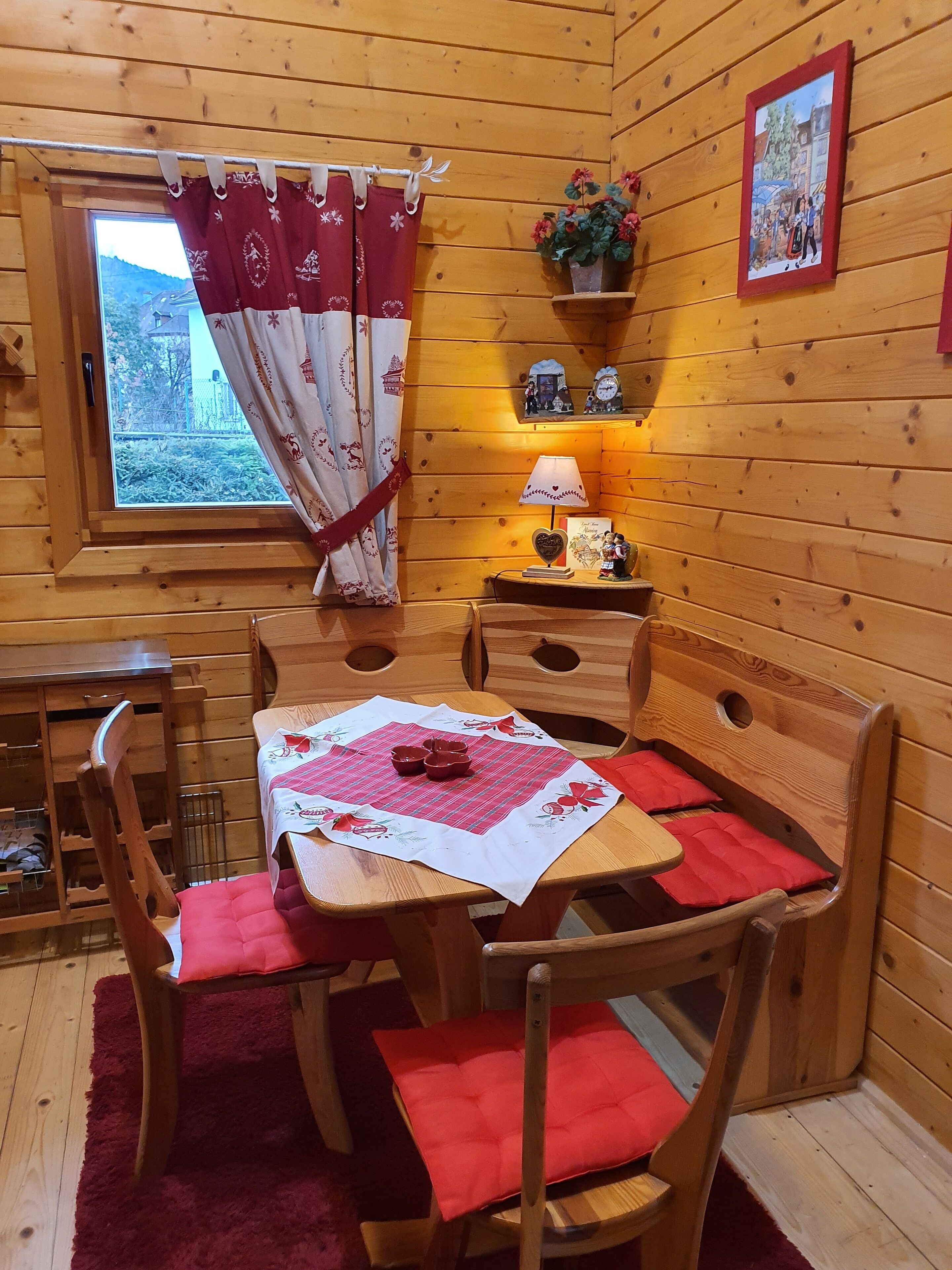 Interior acogedor de una cabaña de madera con una mesa de comedor lista para cuatro personas, una ventana con cortinas, un estante con adornos y una lámpara.