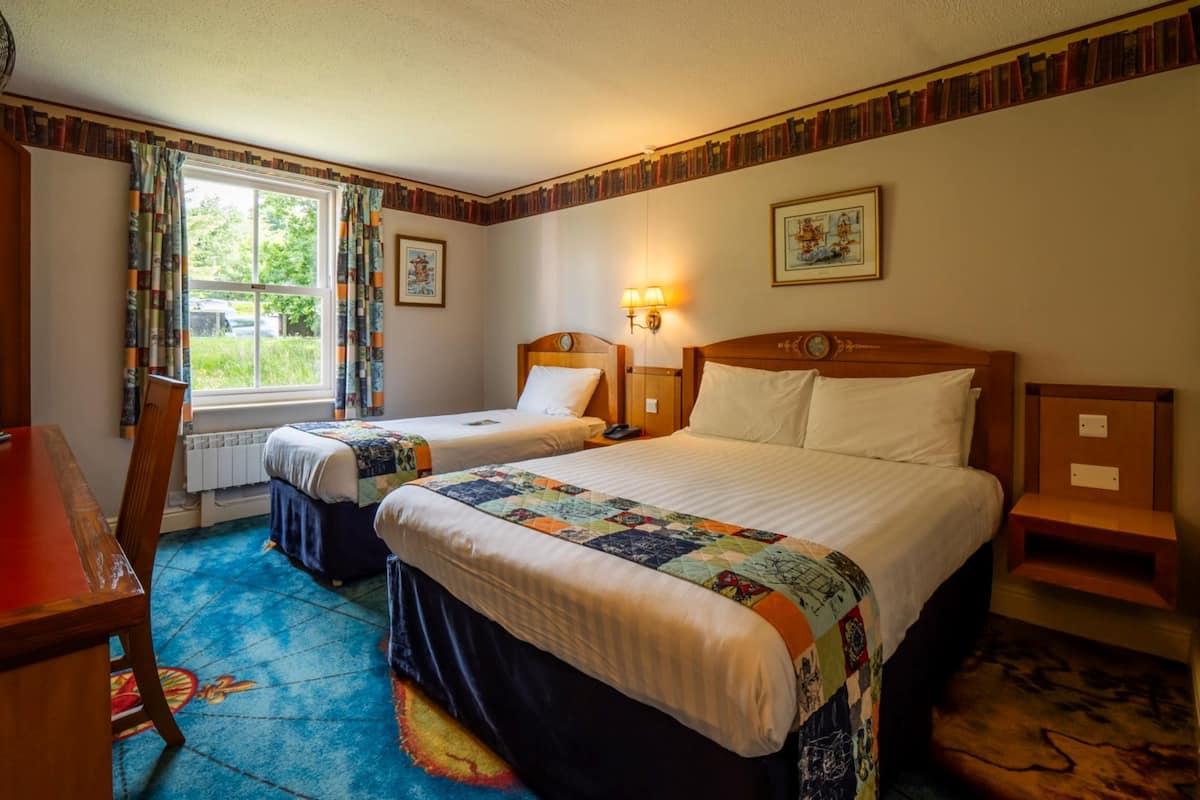 Une chambre d’hôtel avec deux lits, un bureau, une chaise, une fenêtre avec des rideaux, une lampe fixée au mur et un tableau encadré.