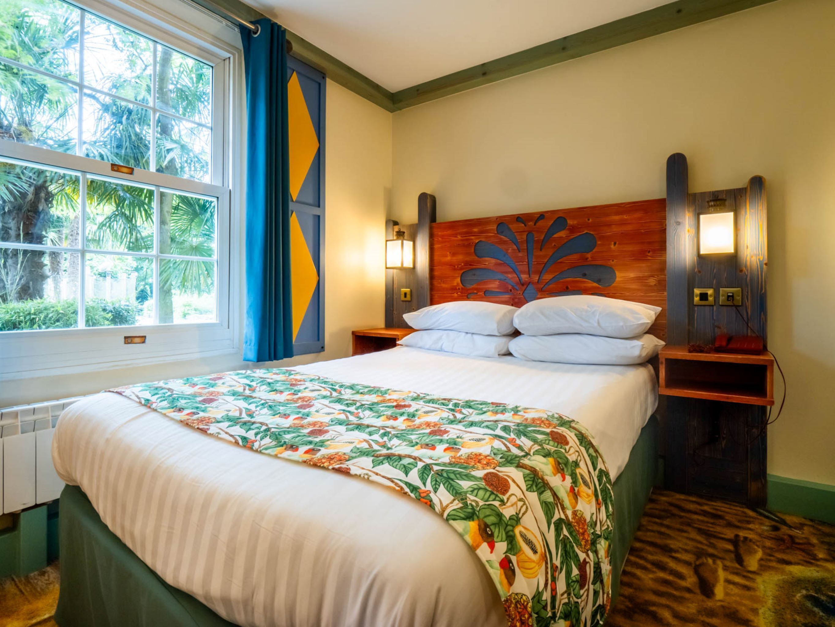 Habitación de hotel con cama, ventana con cortinas, cabecero con diseño decorativo, lámparas de noche y mesita de noche de madera.