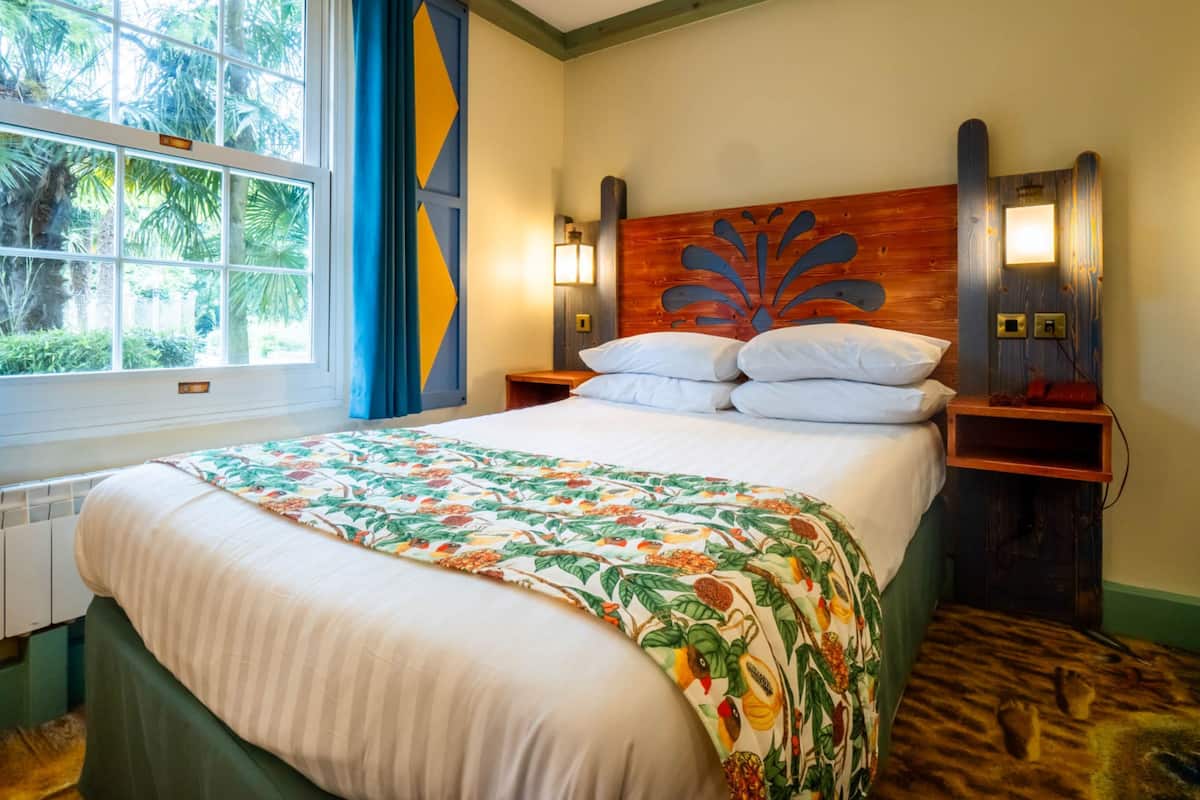 Une chambre d’hôtel comprenant un lit, une fenêtre avec des rideaux, une tête de lit ornée d’un motif décoratif, des lampes de chevet et une table de chevet en bois.