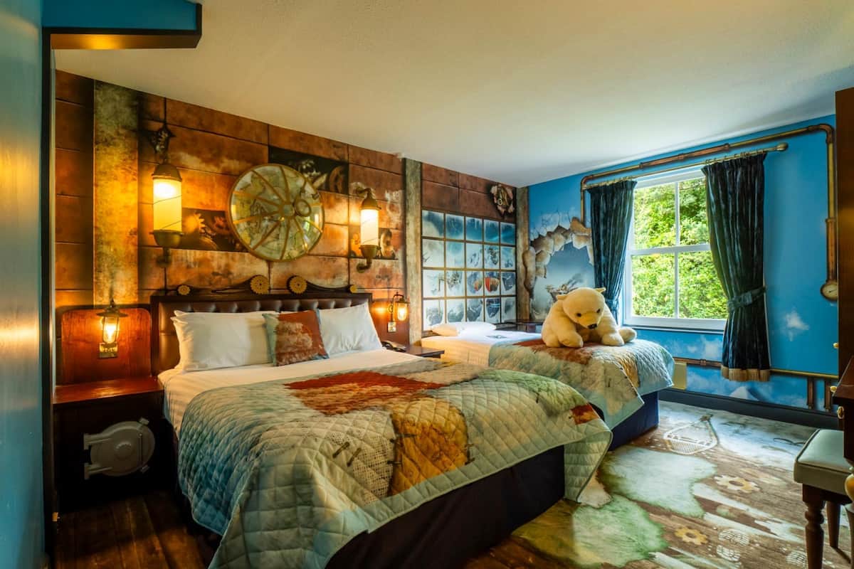 Une chambre avec un grand lit, un plus petit lit, une horloge murale, une fenêtre avec des rideaux et un ours en peluche sur le petit lit.