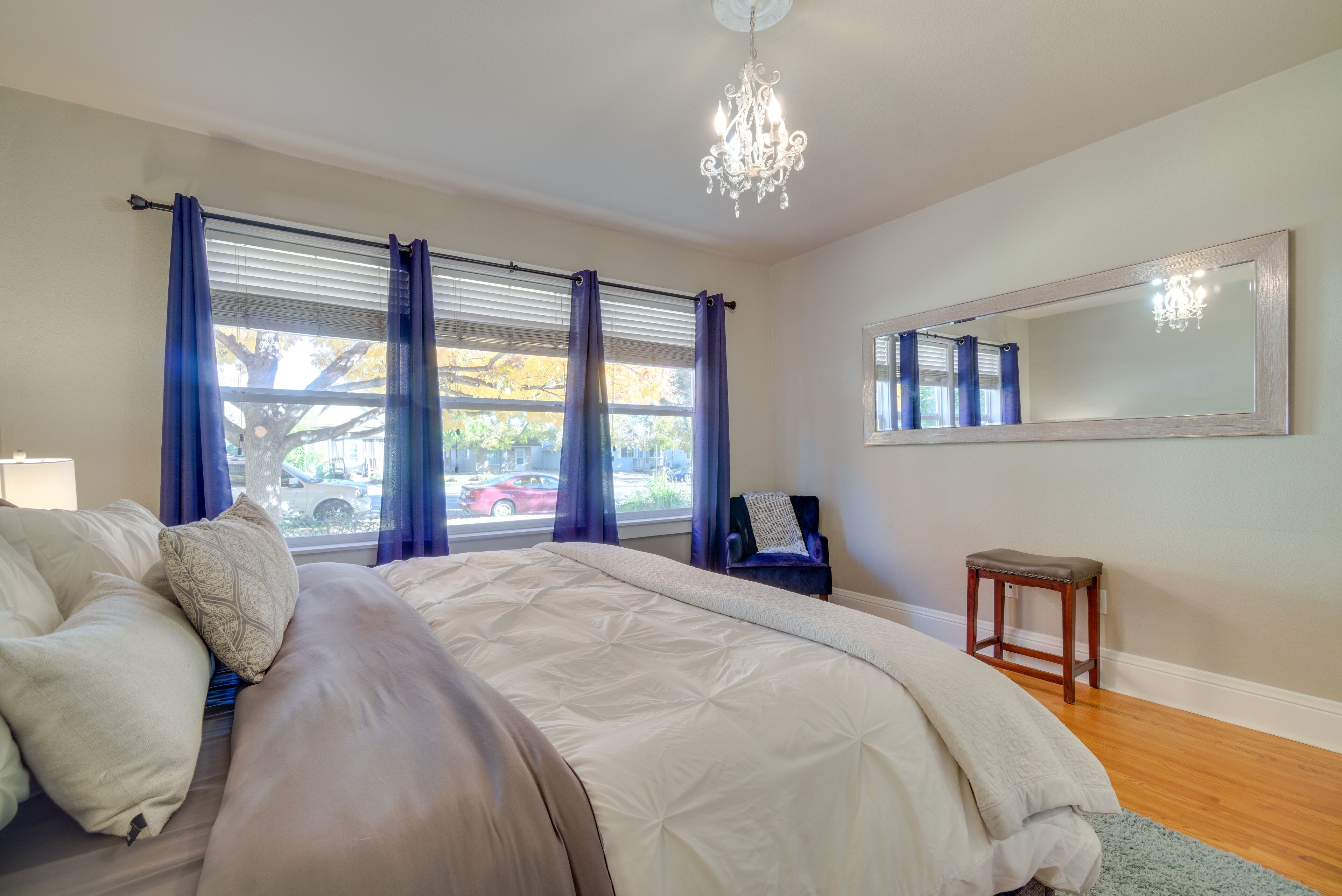 A bedroom with a large bed, a chandelier, a window with curtains, a chair, and a small stool.