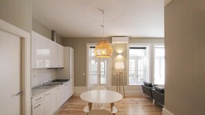 A modern kitchen with white cabinets, a marble countertop, and a round dining table with chairs. There is a floor lamp and a sofa in the adjacent living area. - Duque de Loulé (Porto)