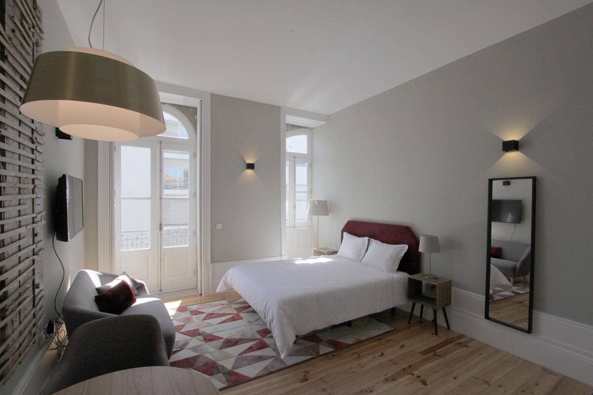 A bedroom with a bed, a chair, a lamp, a mirror, and a geometric-patterned rug.