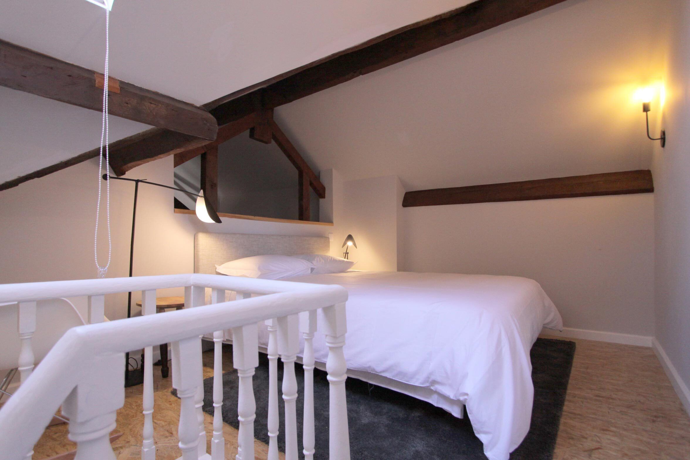 A bedroom with a bed, a white railing, a wall-mounted light, and a wooden ceiling beam.