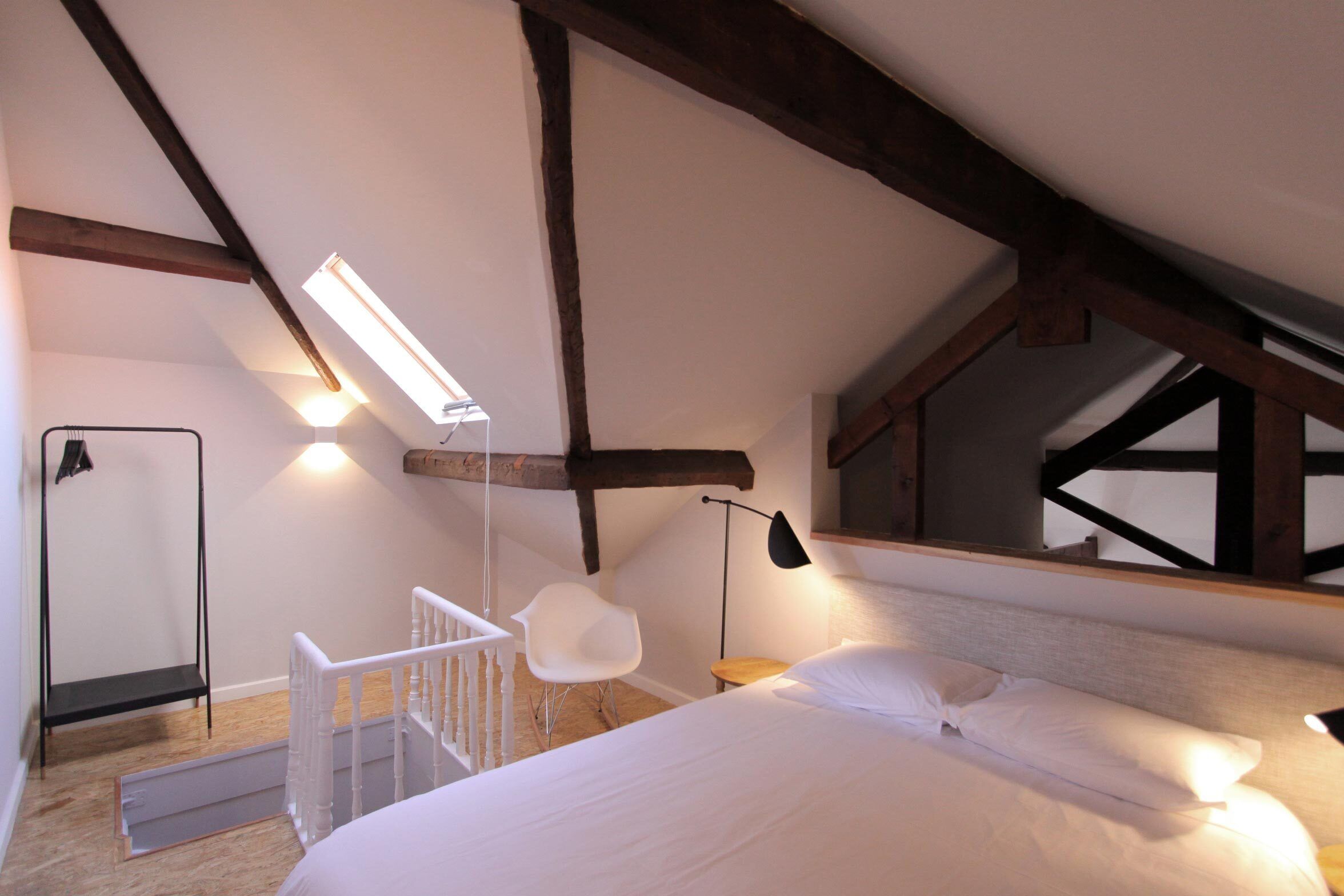 Une chambre avec un plafond en pente, un lit avec une literie blanche, une chaise blanche, une petite table et une lampe sur pied.