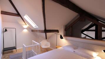 Une chambre avec un plafond en pente, un lit avec une literie blanche, une chaise blanche, une petite table et une lampe sur pied.