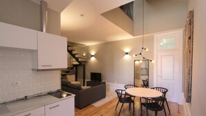A modern kitchen with white cabinets, a marble countertop, and a built-in stove. There is a dining area with a round table and four chairs. A staircase leads to an upper level. - Duque de Loulé (Porto)