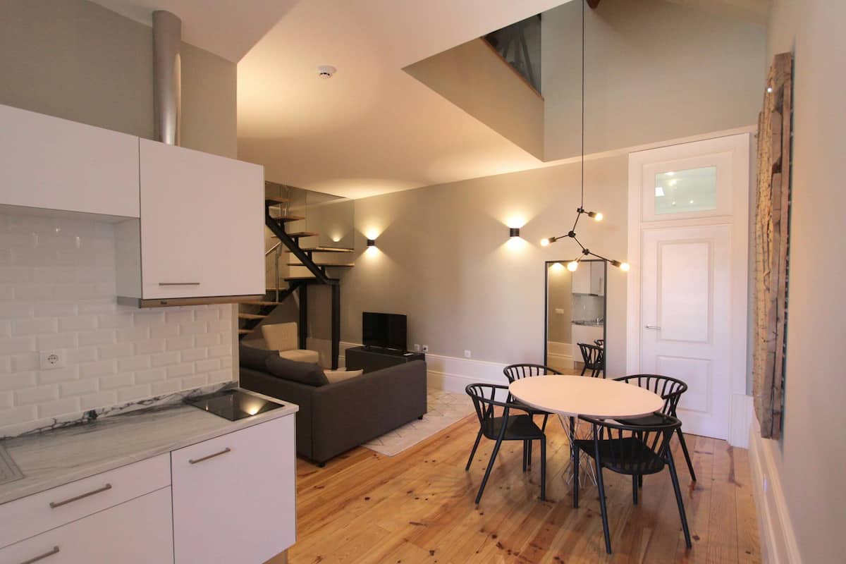 A modern kitchen with white cabinets, a marble countertop, and a built-in stove. There is a dining area with a round table and four chairs. A staircase leads to an upper level.