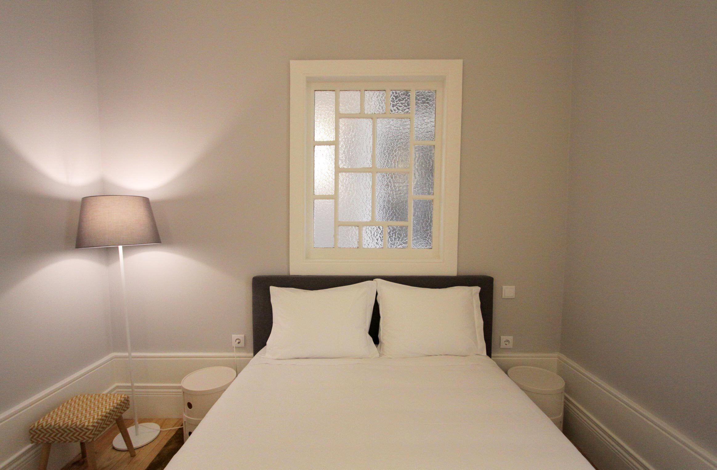 Un dormitorio con cama, mesita de noche, lámpara, ventana y un taburete pequeño.