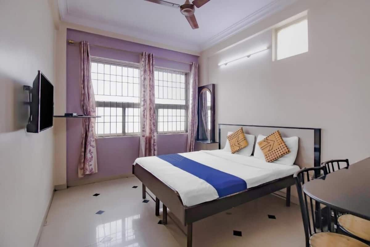 Un dormitorio con una cama, un televisor montado en la pared, un ventilador de techo y una ventana con cortinas.