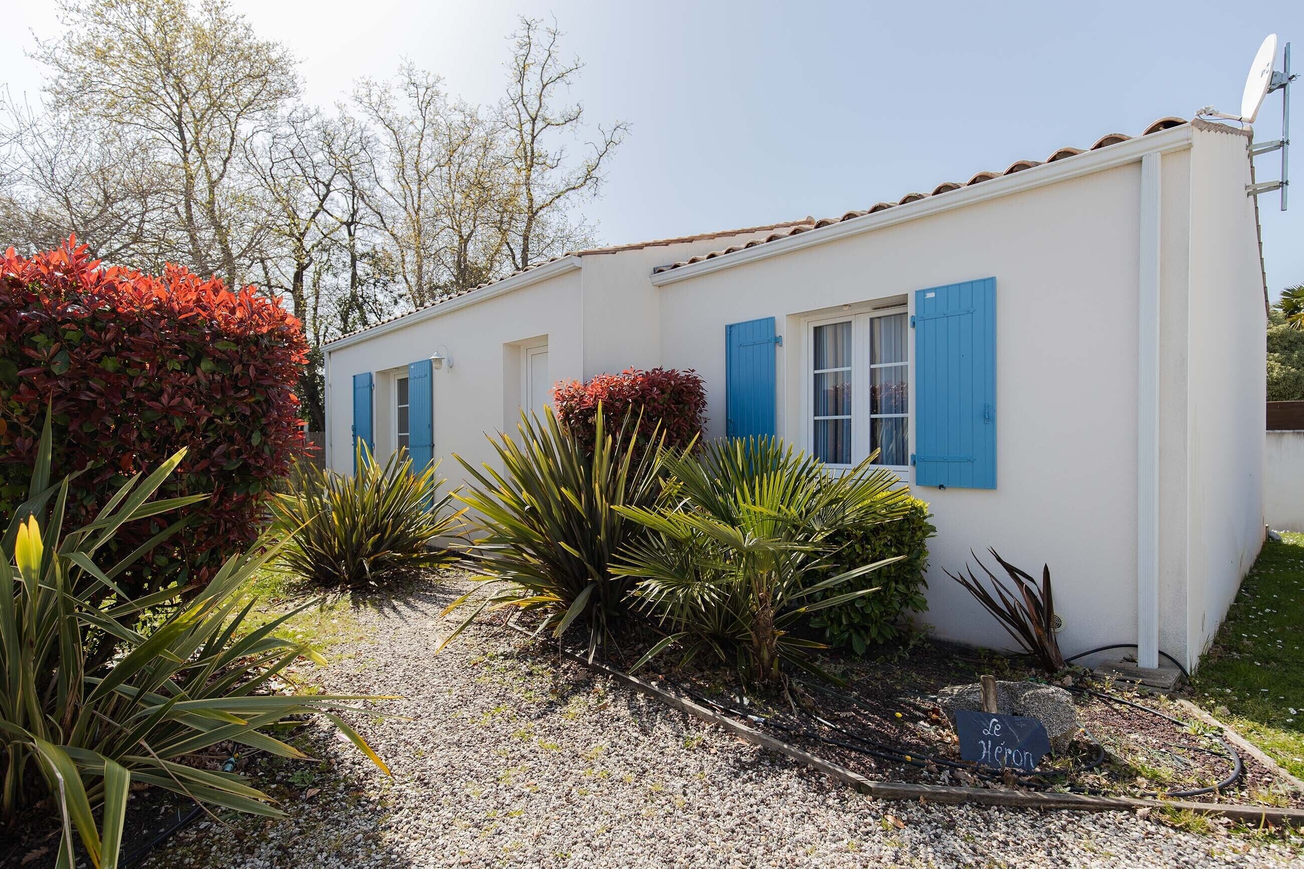 Un bâtiment blanc aux volets bleus, entouré de différentes plantes et bordé d’un chemin de gravier.