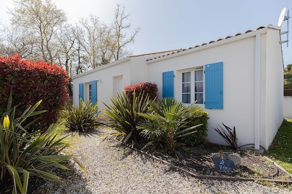 A white building with blue shutters, surrounded by various plants and a gravel path. - Villa in a small village on the island of Oleron (Dolus-d'Oléron)