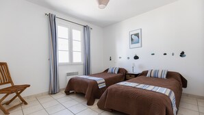 A bedroom with two beds, a wooden chair, a window with curtains, and a wall-mounted lamp.