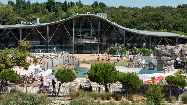 A modern building with a curved roof and large glass windows, surrounded by a pool area with people and sun loungers.