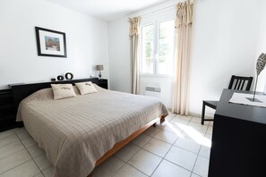 A bedroom with a bed, bedside table, lamp, and a view of the outdoors through a window with curtains. - Villa in a small village on the island of Oleron (Dolus-d'Oléron)