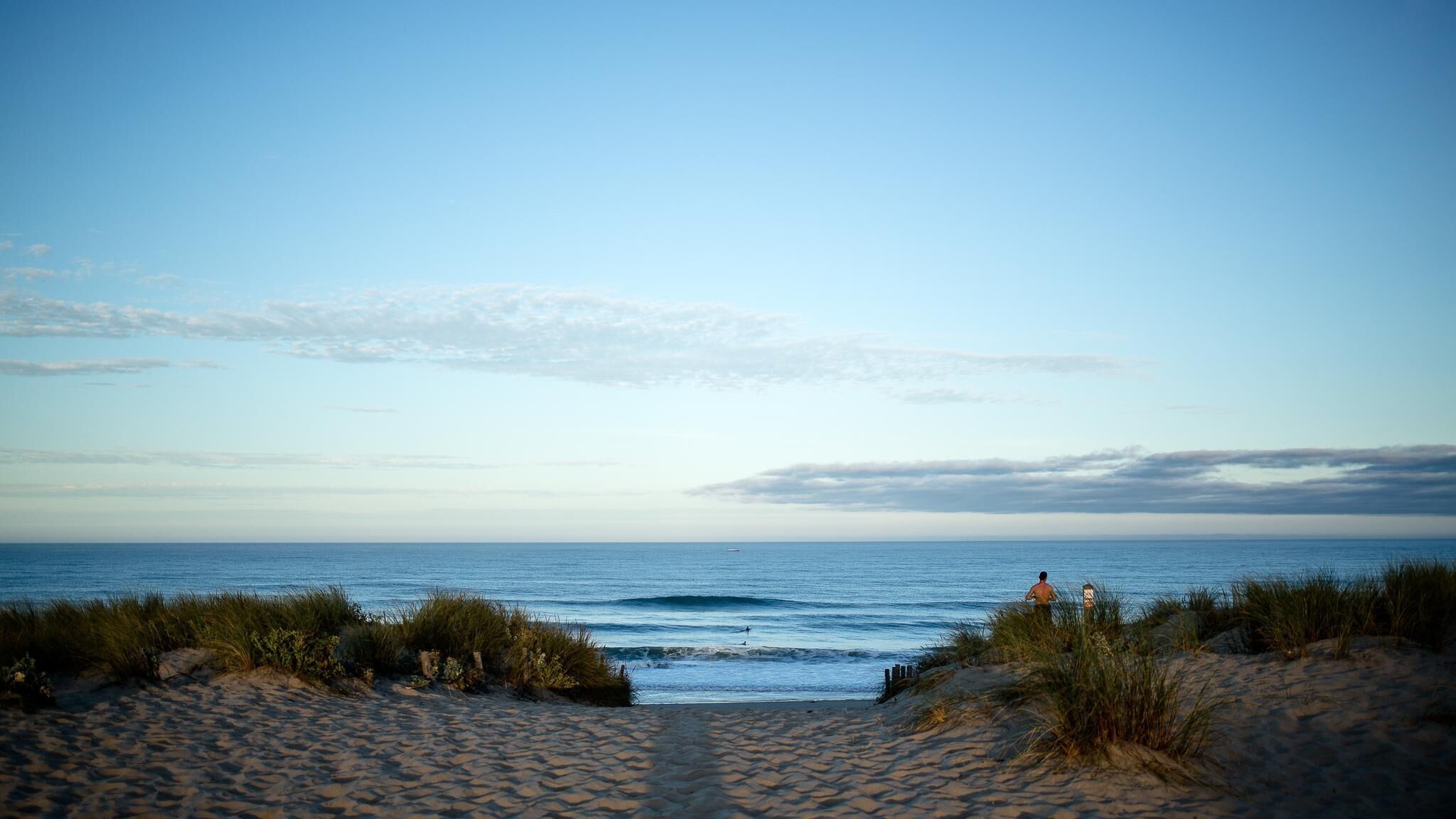 A beach with a clear sky, sandy path leading to the water, and a lifeguard tower in the distance.