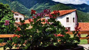 A white building with a red-tiled roof, surrounded by lush greenery and mountains in the background.