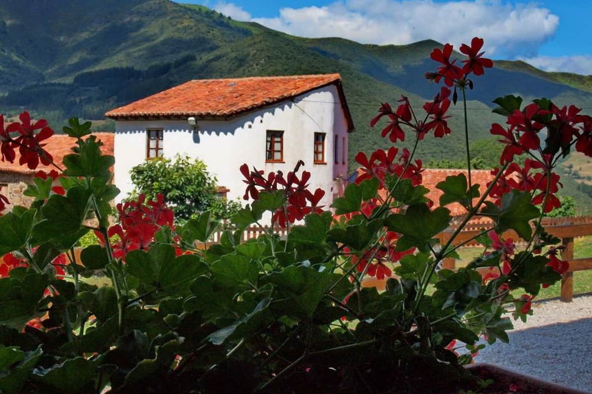Un edificio blanco con techo rojo, rodeado de vegetación y montañas al fondo.