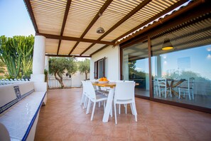 An outdoor patio with white chairs and a wooden table, a covered area with a metal roof, and a view through glass doors to the interior. - Independent villa between Trapani and San Vito Lo Capo, with swimming pool and sea view (Valderice)