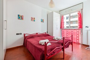Una stanza con un letto in metallo rosso, pareti bianche e una finestra con tende rosse.