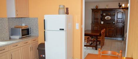 A kitchen with a refrigerator, microwave, and wooden cabinets. There is a trash bin and a dining table with chairs.