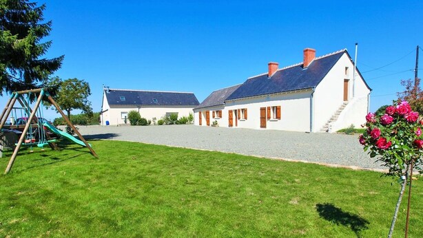 Un bâtiment blanc à deux étages, avec une allée de gravier, une balançoire et un petit jardin agrémenté de fleurs roses.