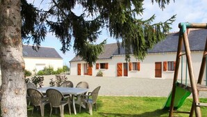 Une terrasse avec une table et des chaises, une balançoire et, au fond, un bâtiment avec une allée gravillonnée.