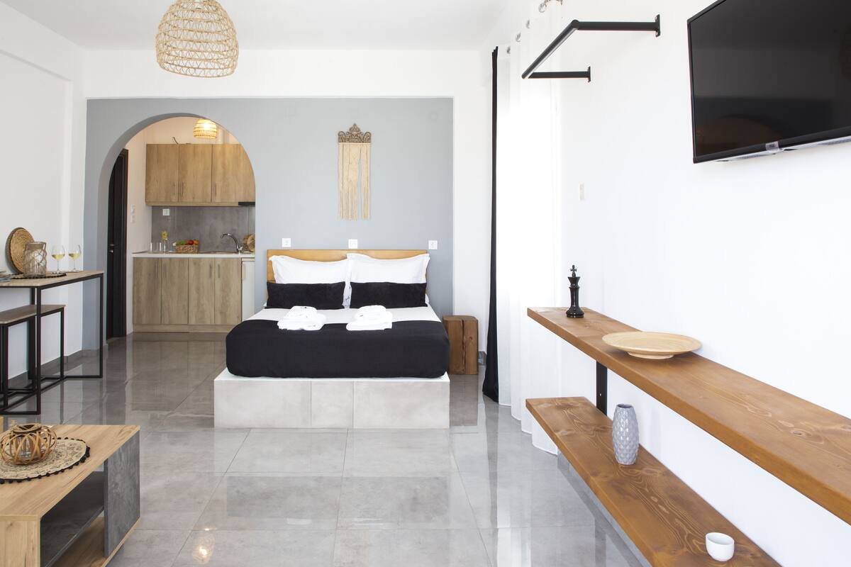 Ein modernes Hotelzimmer mit einem Bett, einer Holzbank, einem kleinen Tisch, einem Fernseher und einer Küchenzeile im Hintergrund.