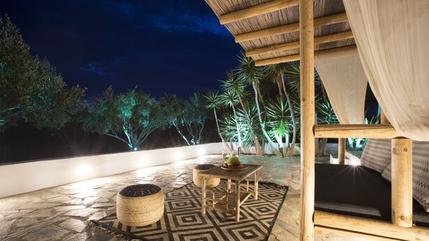 An outdoor patio area with a stone floor, a patterned rug, a wooden table with a plant, and a cushioned seating area under a thatched roof. - Paros King Luxury Apartments (Paros)
