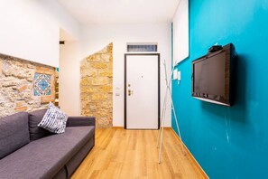 A room with a stone wall, a blue wall, a grey sofa, a white door, and a mounted TV. - Apartment in the heart of the Ballarò market (Palermo)