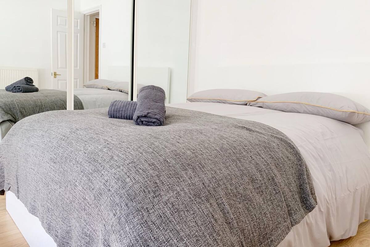 A neatly made bed with a grey blanket and rolled towels, a white headboard, and a white door in the background.