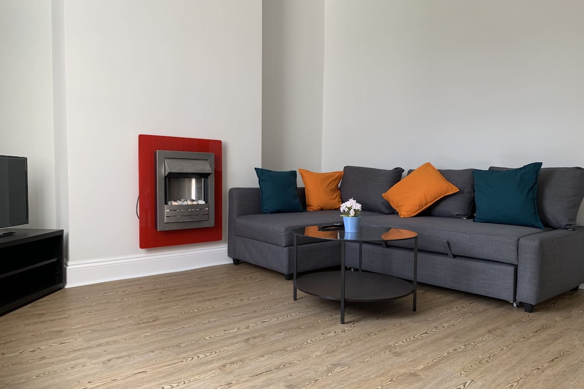 A modern living room with a grey sofa, orange and teal cushions, a small round coffee table, and a red fireplace.