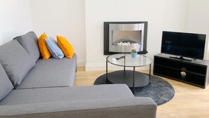 Un salon moderne avec un canapé gris, une table basse en verre, une cheminée et un téléviseur posé sur un meuble noir.