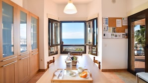 Un comedor con una mesa preparada para dos personas, vista al mar a través de ventanas abiertas y un tablón de anuncios con varios papeles y notas en la pared.