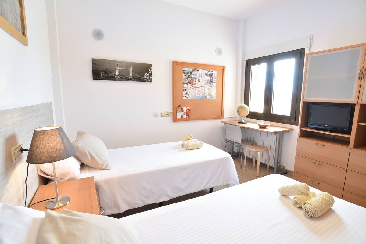 Une chambre d’hôtel avec deux lits, un bureau avec une chaise, une télévision et un tableau d’affichage où sont accrochées des photos et des notes.
