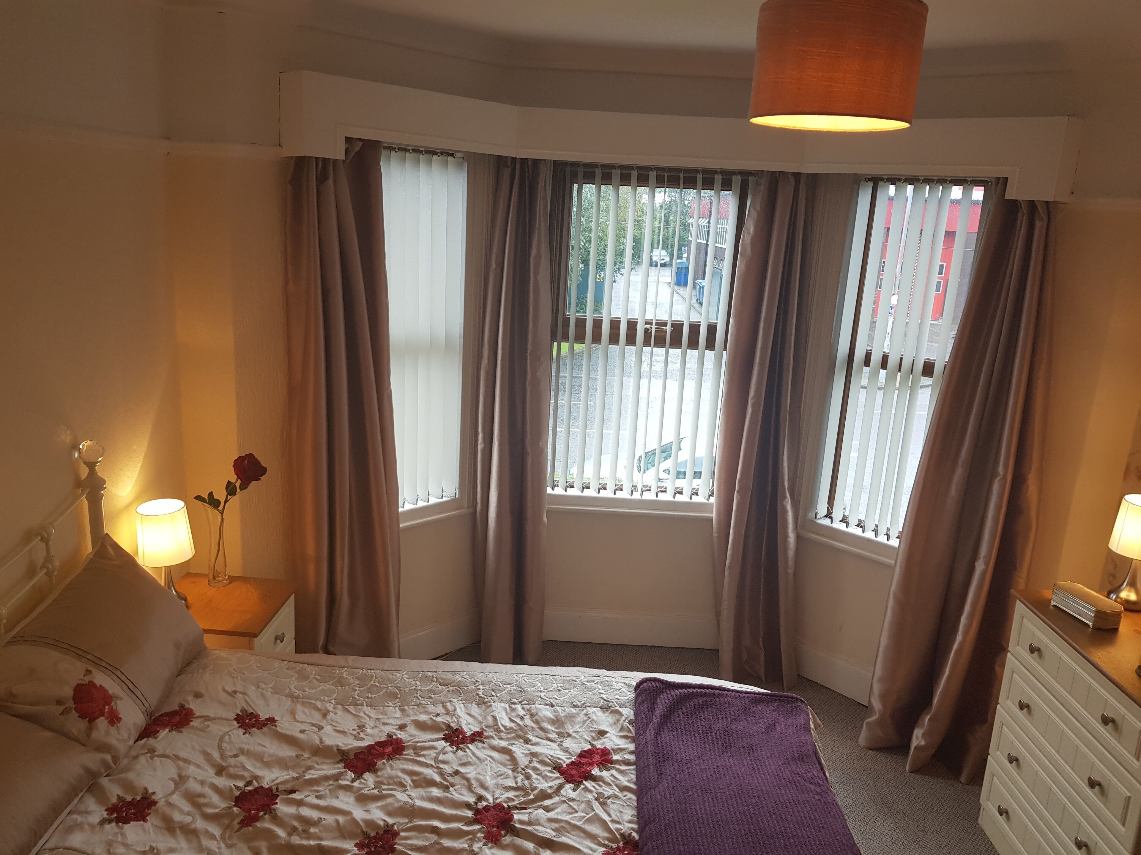A bedroom with a bed, two bedside lamps, a window with curtains, a dresser, and a small table with a lamp and a vase with a single rose.