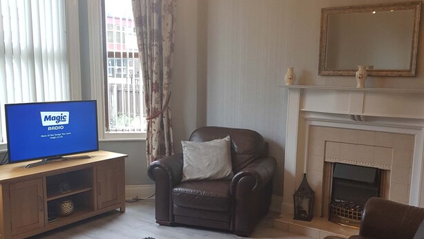 A living room with a leather sofa, a brown armchair, a wooden TV stand with a Magic Radio, a fireplace, and a chandelier. - Delightful Victorian House (Belfast)