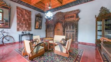 A room with a traditional wooden ceiling, a patterned wall, and a red tiled floor. There is a bicycle, a small table, and a chair with a woven seat.