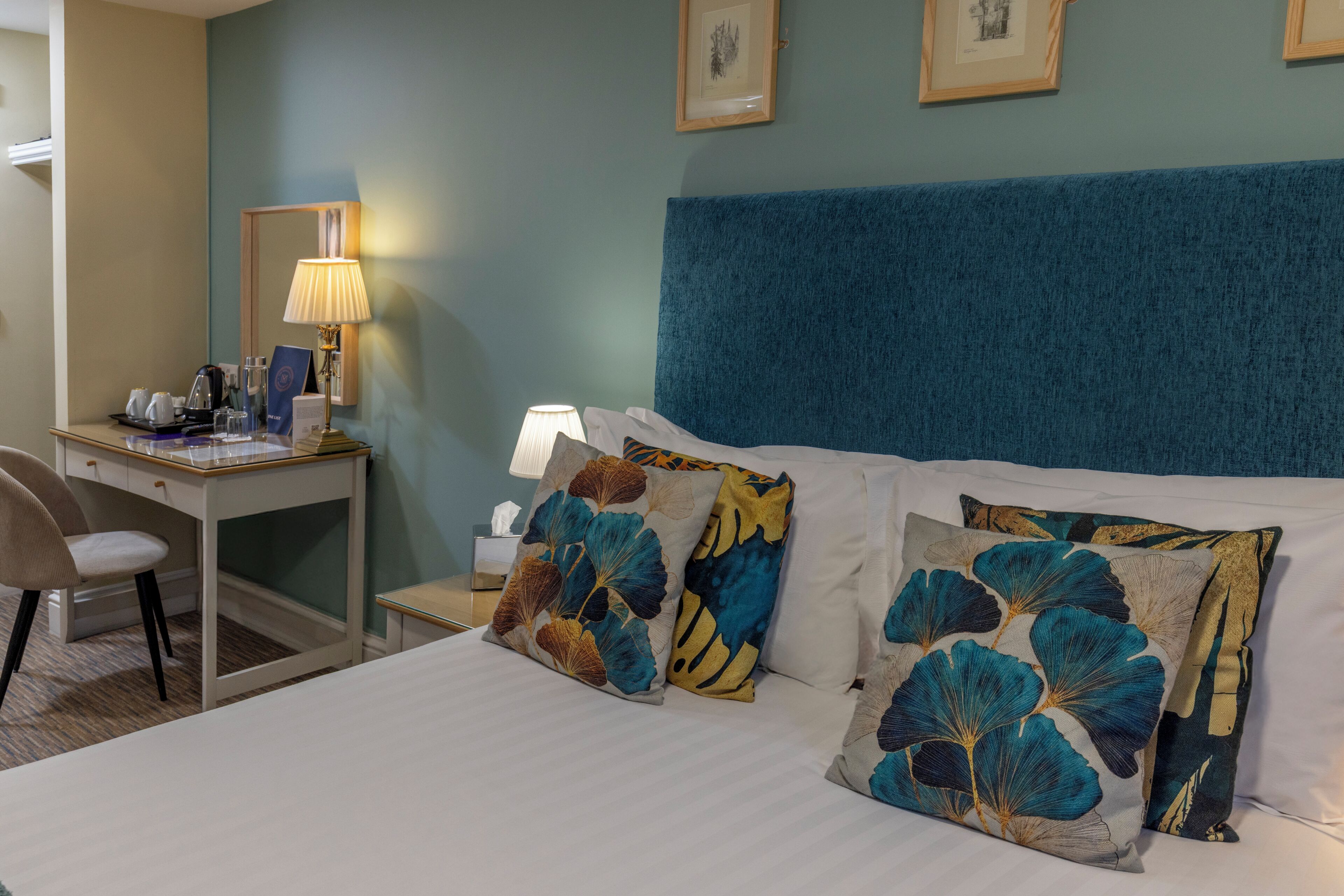 A hotel room with a bed, a desk, a chair, a nightstand, and a lamp.