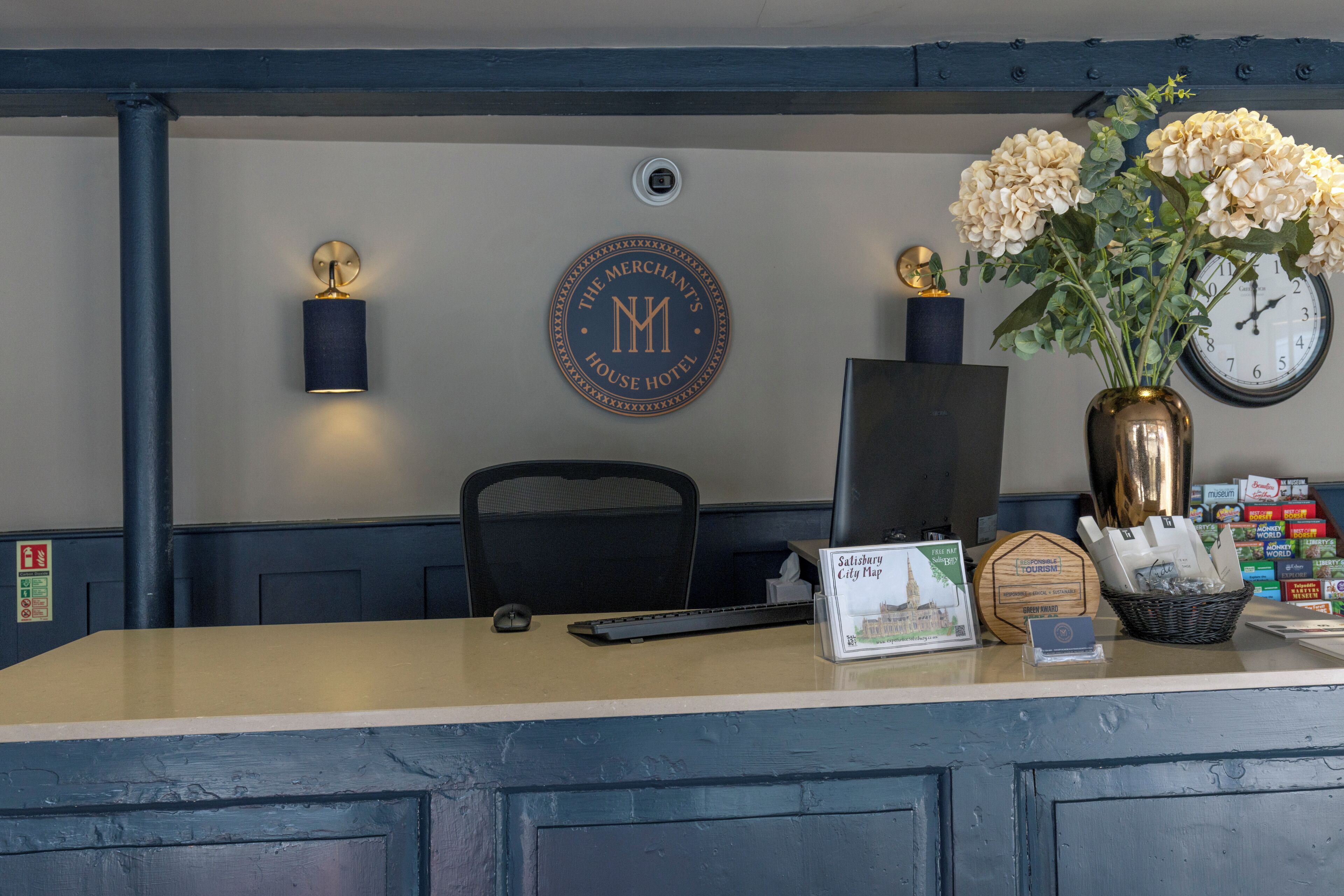 A hotel reception desk with a clock, a framed welcome sign, a vase with flowers, and a computer monitor.