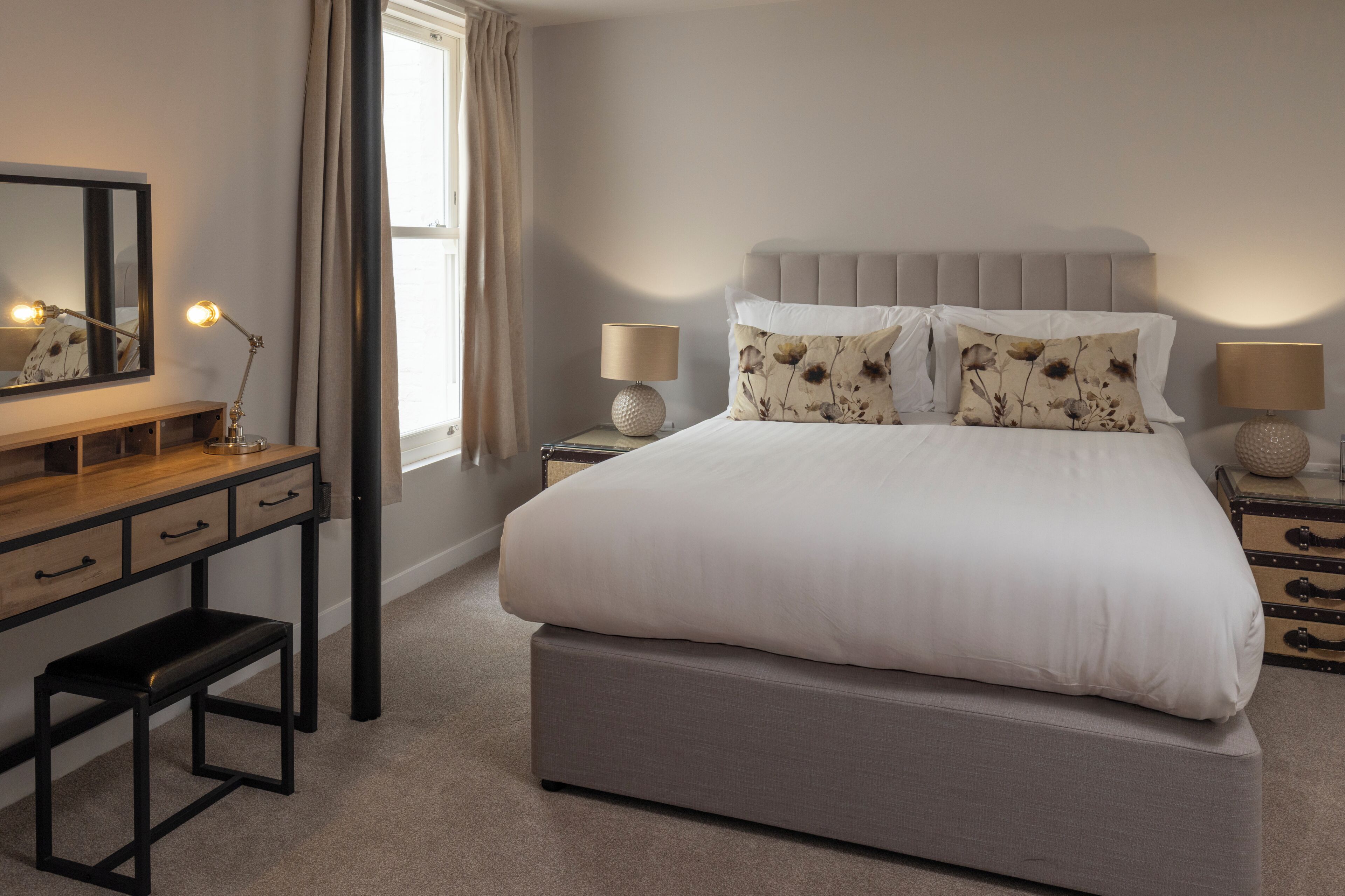 Une chambre à coucher comprenant un lit, des tables de chevet, un bureau, une chaise, un miroir et une fenêtre avec des rideaux.