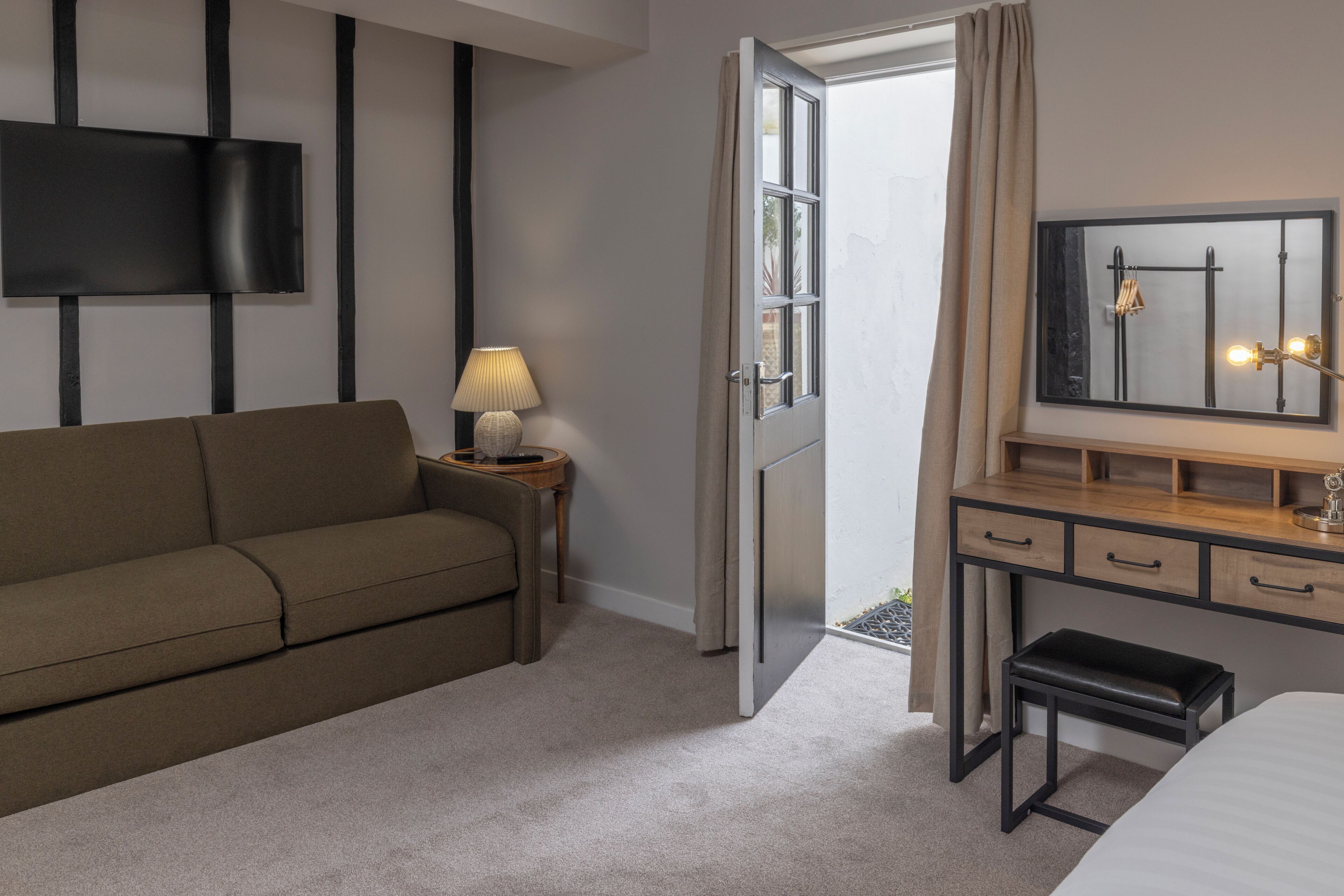 A hotel room with a sofa, a bedside table with a lamp, a dresser with drawers, a mirror, and a door leading to a balcony.