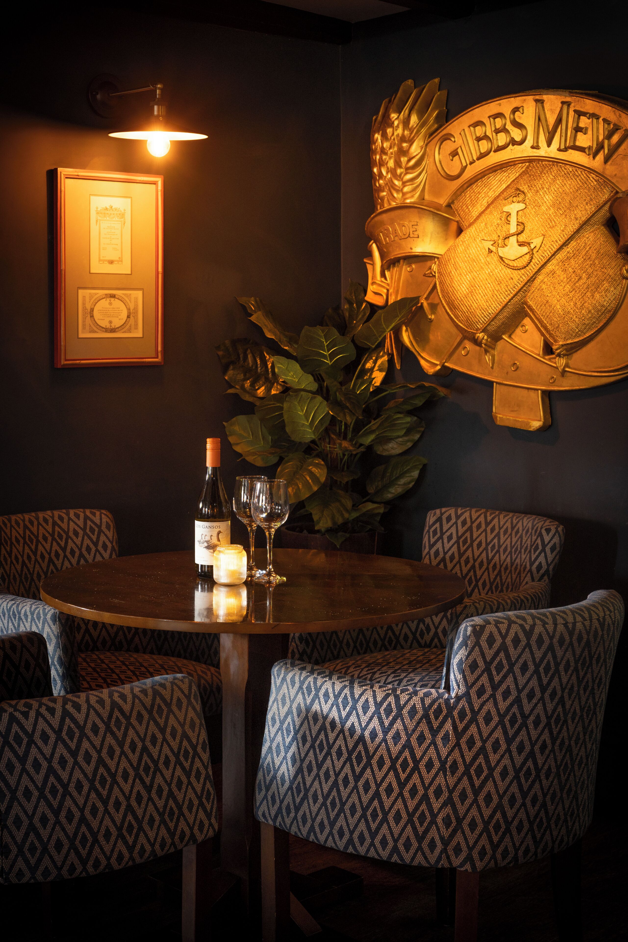 Un coin repas avec une table ronde, des chaises à motifs, une bouteille de vin, des verres, une bougie, un certificat encadré et un grand écusson décoratif portant l’inscription « GIBBS MEW » au mur.